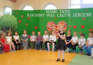 Dziewczynka trzyma w ręku mikrofon, recytuje wiersz, w tle siedzą dzieci w kręgu.
